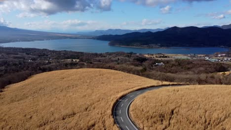 Una-Carretera-Con-Curvas-Que-Atraviesa-Hierba-Dorada-Con-Un-Lago-Y-Montañas-En-La-Distancia,-Vista-Aérea