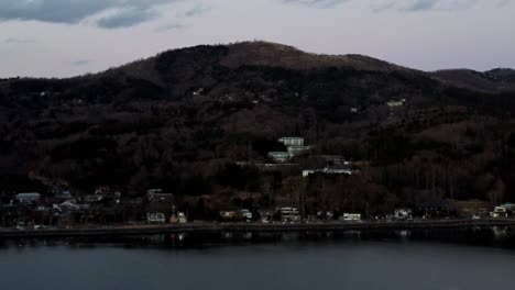 El-Crepúsculo-Desciende-Sobre-Una-Serena-Ciudad-Junto-Al-Lago-Con-Colinas-Boscosas-Y-Un-Claro-Reflejo-En-El-Agua,-Filmada-Con-Poca-Luz.