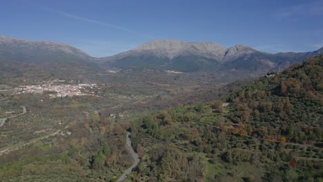 flight-in-a-valley-in-autumn-with-colorful-trees-with-a-road-and-cars-driving,-in-pine-forests-with-a-background-of-granite-mountains-and-a-town-on-a-day-with-blue-sky-in-Avila-Spain