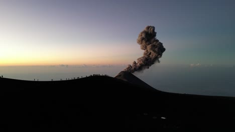 Drone-view-in-Guatemala-flying-over-a-volcano-crater-with-an-erupting-volcano-at-sunrise-and-shadow-of-people-on-the-rim