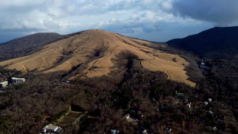 A-vast-barren-hill-with-scattered-homes-at-its-base,-under-a-cloudy-sky,-aerial-view