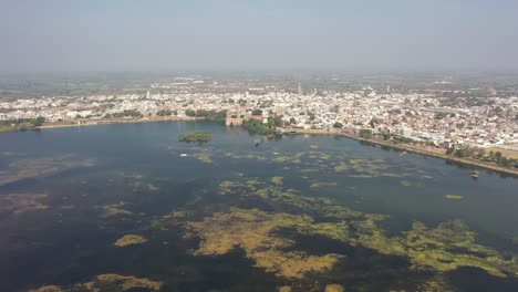 Aerial-drone-view-drone-camera-is-moving-forward-many-birds-are-flying-many-residential-houses-are-also-visible,-Samantsar-Lake,-Halvad-Palace,-Morbi