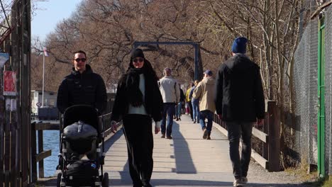 Young-couple-with-stroller-and-others-walk-over-wooden-bridge-in-Stockholm,-sunny-day