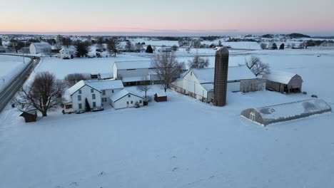 Luftaufnahme-Einer-Schneebedeckten-Farm-In-Der-Abenddämmerung-Mit-Mehreren-Gebäuden-Und-Einem-Silo