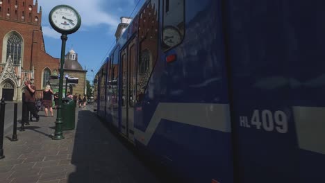 Blauer-Trolley-Fährt-An-Einer-Alten-Eisenbahnuhr-An-Einer-Belebten-Straße-In-Krakau,-Polen-Vorbei
