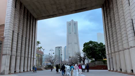 People-walking-through-a-large-concrete-gate-towards-a-tall-skyscraper,-overcast-sky,-urban-setting,-daytime,-in-Yokohama,-Japan