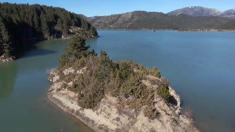 Vista-Aérea-De-La-órbita-De-Un-Islote-De-Lago-De-Montaña-Fondo-De-Picos-De-Día-Soleado-De-Invierno