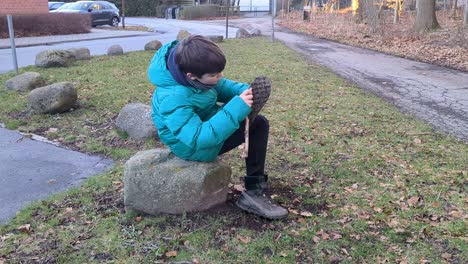 Junge-Sitzt-Auf-Einem-Stein-Und-Putzt-Seine-Schmutzigen-Stiefel-Mit-Einem-Stock