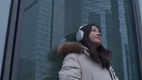 Young-asian-girl-listens-to-music-in-headphones-outdoors,-leaning-against-building-with-skyscraper-reflection-in-window