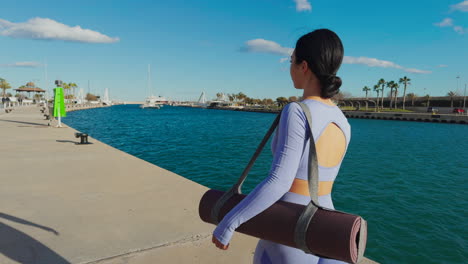 Brünette-Frau-Zu-Fuß-Auf-Der-Promenade-Mit-Yoga-Matte-Und-Sportbekleidung-An-Einem-Sonnigen-Tag,-Rückseite