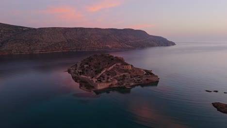 Aerial-drone-pullback-at-sunset-above-Spinalonga-island-fortress-ruins-below-soft-glowing-clouds