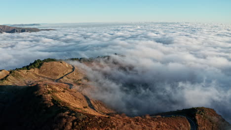 A-winding-road-on-a-mountain-above-the-clouds-at-sunrise,-aerial-view