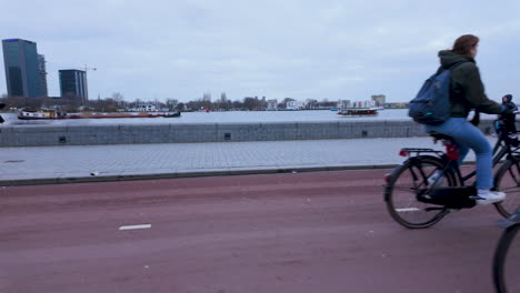 Ein-Paar-Fährt-Zusammen-Fahrrad-Und-Zeigt-Damit-Die-Ungezwungene-Und-Intime-Art-Des-Wichtigsten-Fortbewegungsmittels-In-Amsterdam-Vor-Der-Kulisse-Der-Uferpromenade-Der-Stadt.