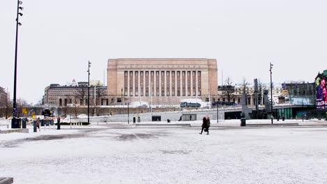 Snowy-Helsinki-square-with-Parliament-House,-people-walking,-overcast-winter-day,-distant-view