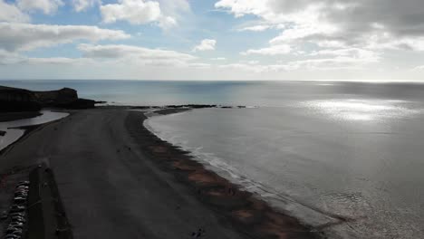 Toma-Aérea-Descendente-Sobre-La-Playa-De-Budleigh-Salterton,-Devon,-Inglaterra,-Con-El-Sol-Brillando-En-El-Mar-En-Calma.