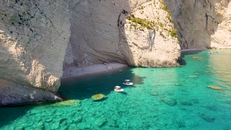 Oasi-beach-with-boats-near-keri-caves,-zakynthos,-crystal-clear-waters-and-steep-cliffs,-sunny-day,-aerial-view