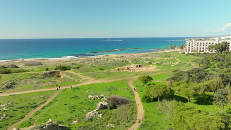 The-Tombs-of-the-Kings-site-is-seen-from-a-distance,-with-tourists-walking-among-the-ruins,-set-against-the-deep-blue-sea-and-green-landscape,-an-emblem-of-Cyprus's-rich-archaeological-heritage