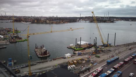 Wide-view-of-cranes-and-buses-at-construction-site-by-water-in-Stockholm