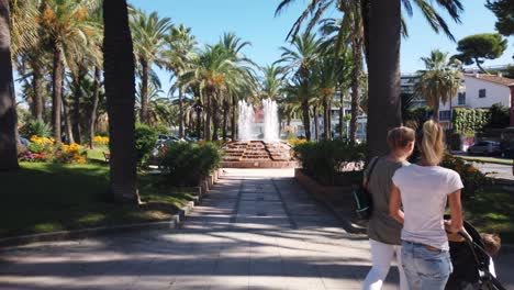 Women-with-stroller-walk-in-park-by-fountain,-French-riviera,-tilt-up