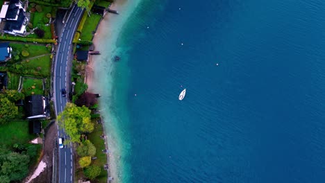 Aerial-view-of-a-road-and-small,-white-sailboat-anchored-in-a-turquoise-bay-with-white-sand-beaches-and-palm-trees,-surrounded-by-lush-green