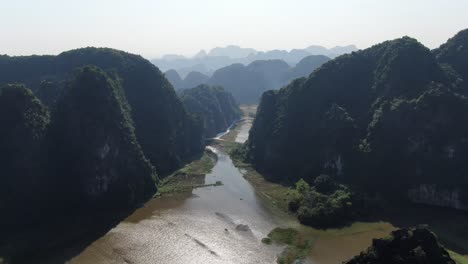 Drone-aerial-view-in-Vietnam-flying-through-rocky-mountains-covered-with-green-trees-over-a-wide-brown-river-in-Ninh-Binh-on-a-sunny-day