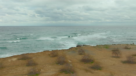 Aerial---tranquil-scene-along-the-coast-of-Cyprus,-where-the-gentle-waves-of-the-Mediterranean-Sea-wash-upon-a-rugged-shoreline-dotted-with-sparse-vegetation-under-a-cloudy-sky