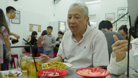 Älterer-Asiatischer-Mann-Isst-Nudeln-Am-Tisch-Im-Restaurant-Mit-Leuten-Um-Ihn-Herum