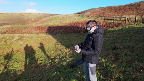 Young-boy-and-his-mother-outside-in-the-countryside,-flying-his-FPV-Drone-with-a-vitual-reality-headset-and-practicing-his-skills-sport