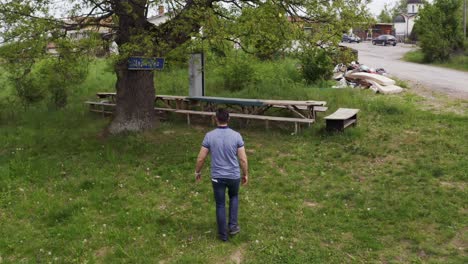 Follow-through-drone-shot-following-a-tourist-walking-towards-a-tree-and-the-benches-located-in-the-village-of-Tsarichina-Hole-in-Bulgaria