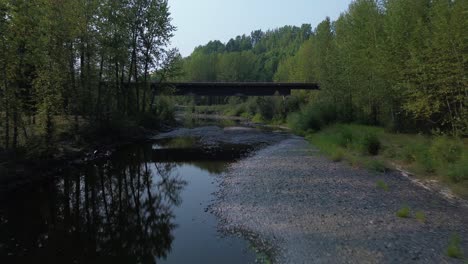 Low-Flying-Drone-Along-the-Bulkley-River-with-a-Low-Bridge-Crossing-and-Alpine-Trees-Either-Side-in-a-Natural-Environment-near-Houston,-Northwood-Picnic-Site,-Canada