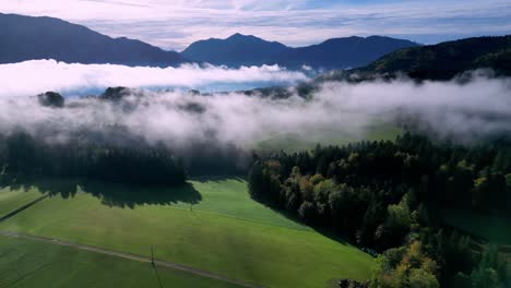 Luftaufnahme-Eines-üppigen-Grünen-Tals-Voller-Nebel,-Durch-Dessen-Mitte-Sich-Ein-Gewundener-See-Schlängelt-Und-In-Der-Ferne-Schneebedeckte-Berge-Aufragen