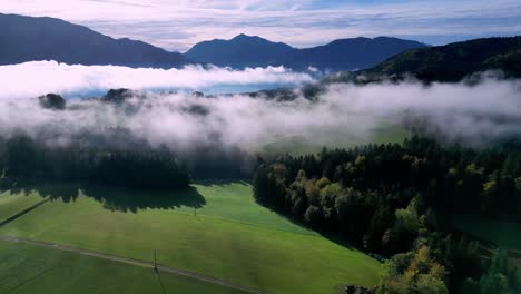 Vista-Aérea-De-Un-Exuberante-Valle-Verde-Lleno-De-Niebla,-Con-Un-Lago-Sinuoso-Que-Serpentea-Por-El-Centro-Y-Montañas-Cubiertas-De-Nieve-Que-Se-Elevan-En-La-Distancia.