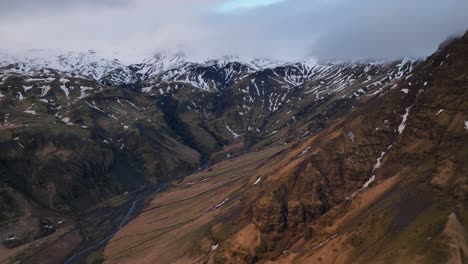 Aerial-landscape-view-of-a-river-flowing-in-a-mountain-valley,-coming-down-from-melting-snow,-in-Iceland,-on-a-cloudy-day