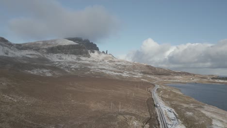 Isle-of-Skye,-Scotland,-coastline-near-with-view-of-snowy-peaks-of-the-Old-Man-of-Storr-mountains-under-a-blue-sky,-and-a-road-by-the-shoreline