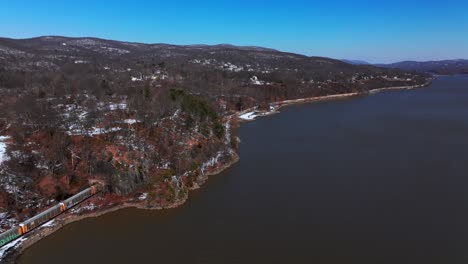 Eine-Luftaufnahme-Eines-Langen-Güterzuges,-Der-An-Einem-Sonnigen-Tag-Mit-Klarem-Blauen-Himmel-Entlang-Eines-Stillen-Hudson-River-Fährt