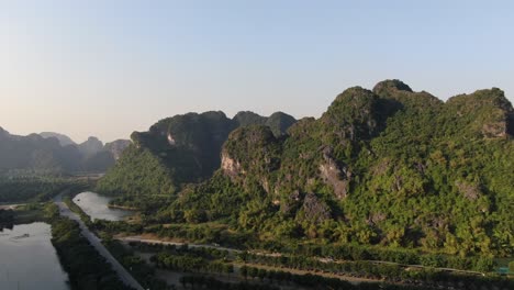 Drone-aerial-view-in-Vietnam-circling-around-a-valley-showing-rocky-mountains-covered-with-green-trees-over-a-river-and-a-road-in-Ninh-Binh-on-a-sunny-day
