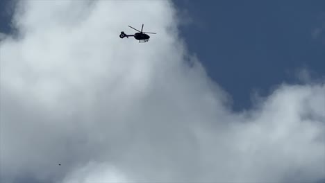 Cámara-Lenta-De-Un-Helicóptero-Volando-Hacia-El-Cuadro-Desde-Arriba-A-La-Izquierda-Con-Nubes-Hinchadas-Detrás-Y-Cielo-Azul-Y-Salidas-Del-Cuadro-A-La-Izquierda.