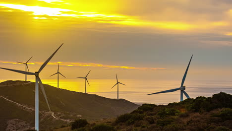 Rotating-Windmills-Closeup-Time-Lapse-Panoramic-Landscape,-Sunset-Clouds-Moving-with-green-Hills,-sun-rays-through-renewable-energy-source