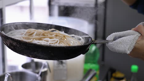 El-Chef-Vierte-Un-Cucharón-De-Salsa-Oscura-Sobre-Fideos-De-Arroz-En-Una-Sartén,-Una-Sartén-Llena-De-Palitos-De-Arroz-Humeantes-Se-Rocía-Con-Salsa-De-Soja,-Cámara-Lenta-De-Cerca-4k