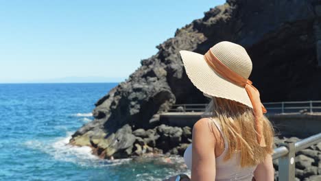 Classy-summertime-blonde-woman-with-hat-enjoys-Atlantic-weather,-back-view