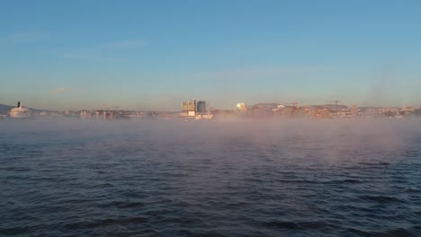 Aerial-Low-Flying-Over-Waters-With-Floating-Fog-Rising-To-Reveal-Bjorvika-In-Sentrum-Borough-of-Oslo,-Norway