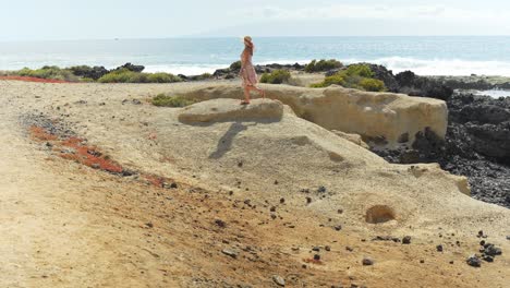 Woman-wearing-floral-dress-walking-over-the-boulders-near-the-beach,-static-slow-motion