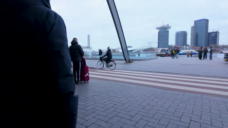 A-bustling-urban-scene-outside-a-modern-transportation-hub,-with-pedestrians,-a-cyclist,-and-a-worker-in-a-high-visibility-jacket,-with-the-city's-skyline-in-the-background