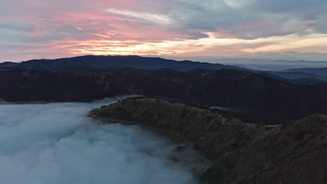 Misty-mountain-range-at-sunset-with-clouds-enveloping-valleys,-aerial-view