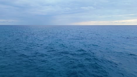 Fast-low-aerial-dolly-out-to-deep-blue-ocean-water-with-storm-cloud-rain-on-horizon