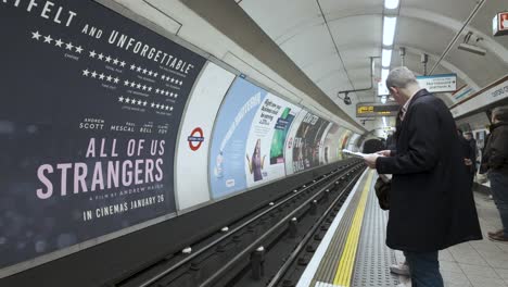 Man-waiting-on-the-Bakerloo-Line-platform-at-Oxford-Street-station-in-London,-adverts-on-wall,-subdued-lighting