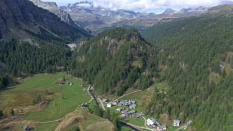 Lago-di-devero,-alpe-devero-with-rustic-houses,-green-fields,-and-surrounding-forests-in-daylight,-aerial-view