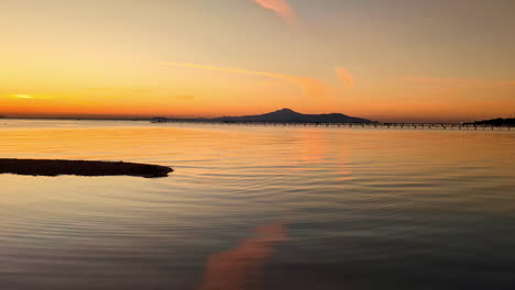 Aerial-Sunset-landscape-above-African-beach-coastline,-volcano-hills-gradient,-orange-golden-light-blue-vibrant-skyline-over-reflected-calm-still-sea-water