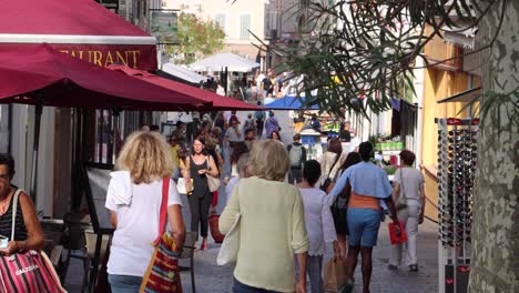People-walk-by-stores-and-restaurants-in-Antibes-in-summertime,-static