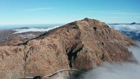Un-Pico-De-Montaña-Escarpado-Sobre-Las-Nubes-Con-Cielos-Azules-Claros,-Vista-Aérea